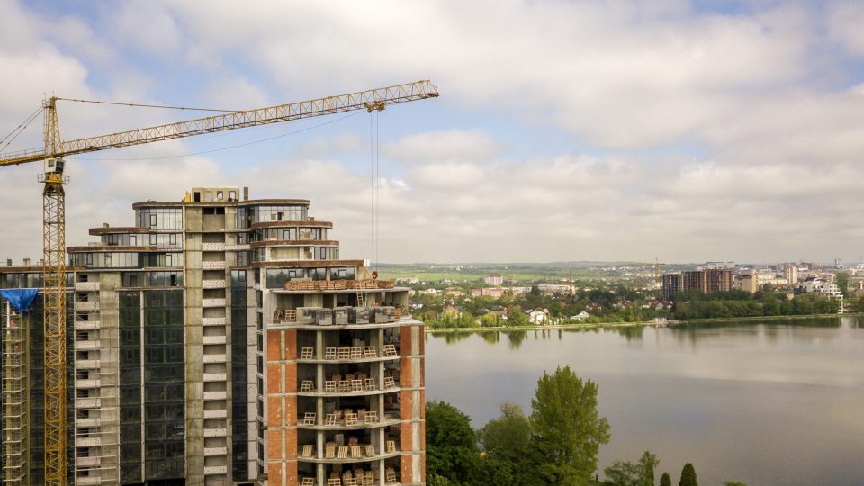 Apartment or office tall building under construction. Brick walls, glass windows, scaffolding and concrete support pillars. Tower crane on bright blue sky copy space background.