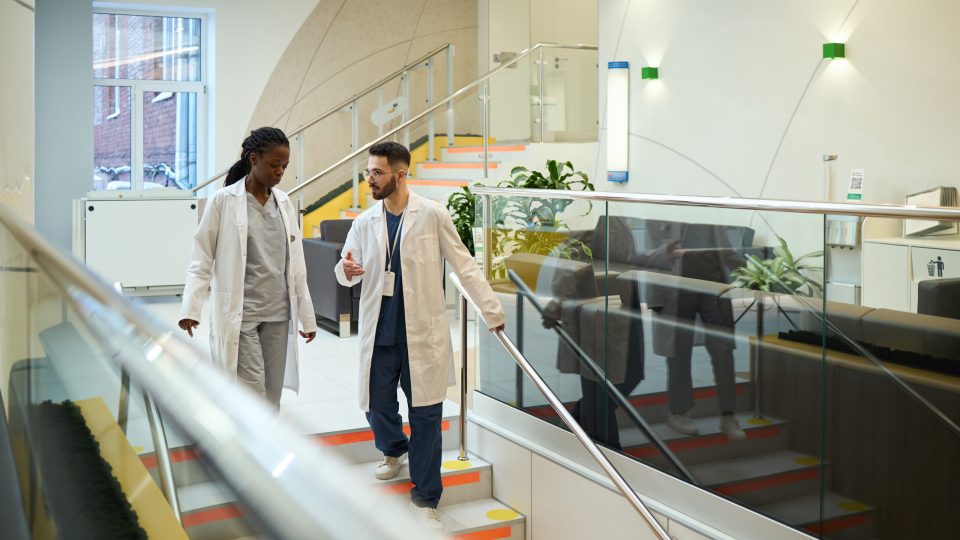 Two medical professionals conversing while walking down stairs in hospital setting, wearing lab coats, focusing on patient care and treatment options